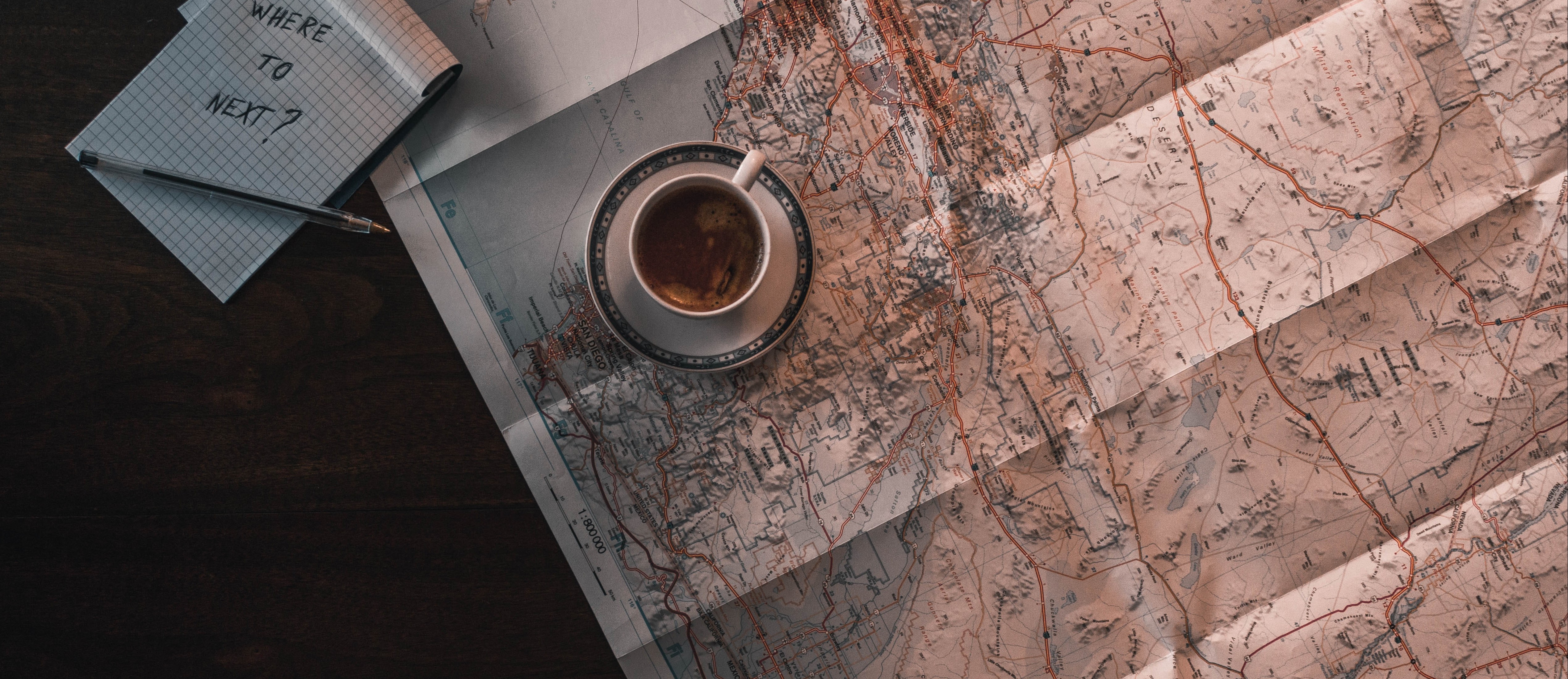 Top-down shot of full coffee cup sitting on paper map. Notebook next to the coffee cup reads: 'Where to next?' 